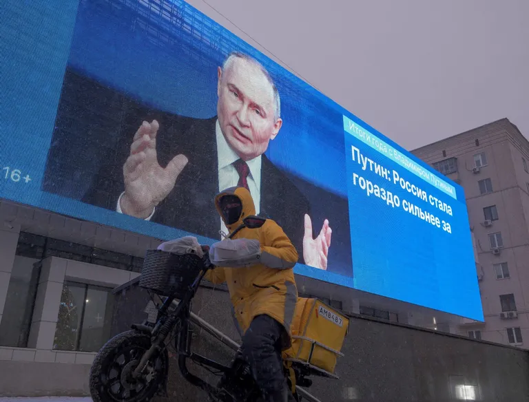Человек в форме службы доставки на электровелосипеде на фоне большого экрана. На экране изображен Владимир Путин и выведена цитата «Путин: Россия стала гораздо сильнее за».