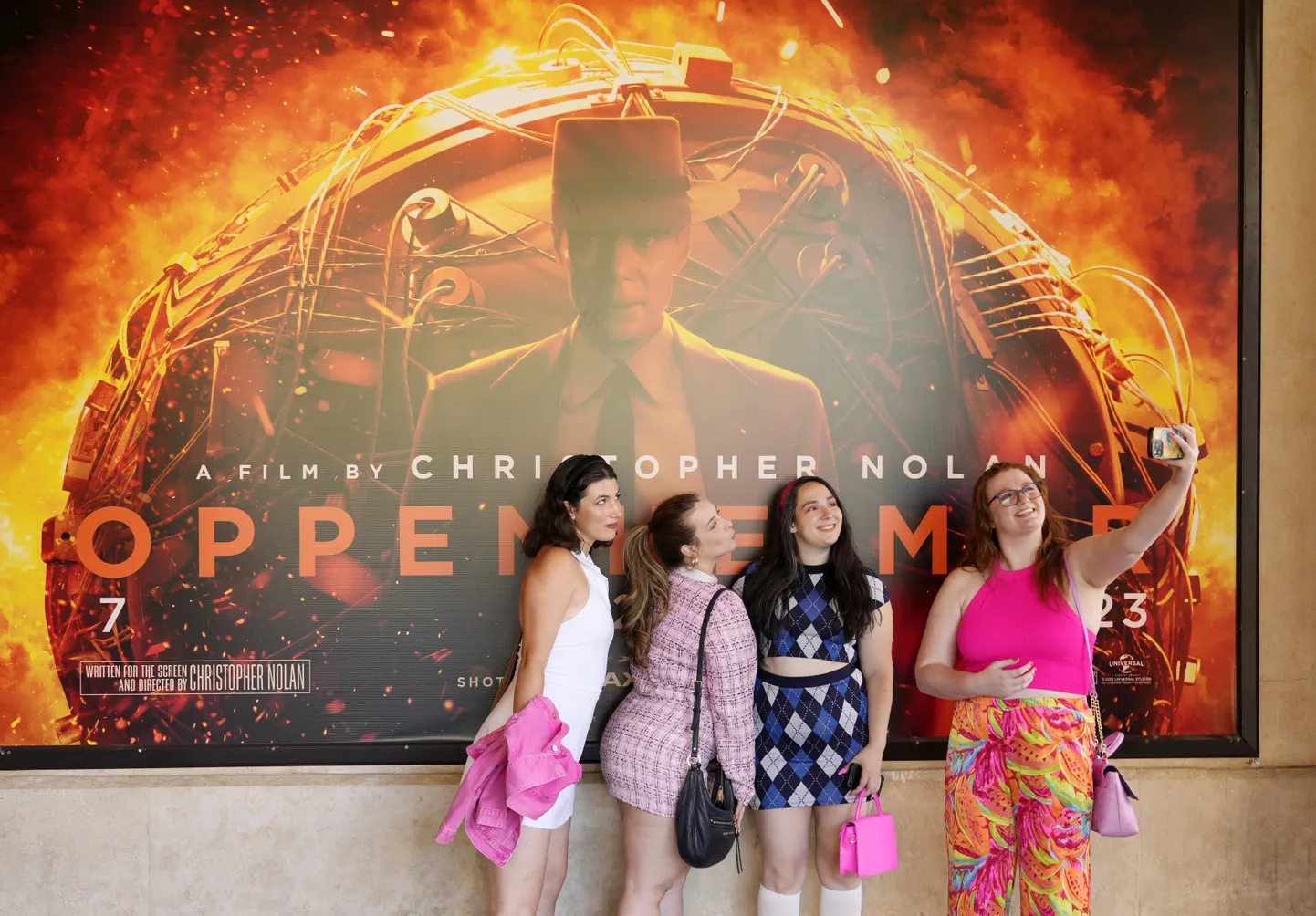 Roosades riietes sõpruskond poseerimas filmi «Oppenheimer» postri taustal