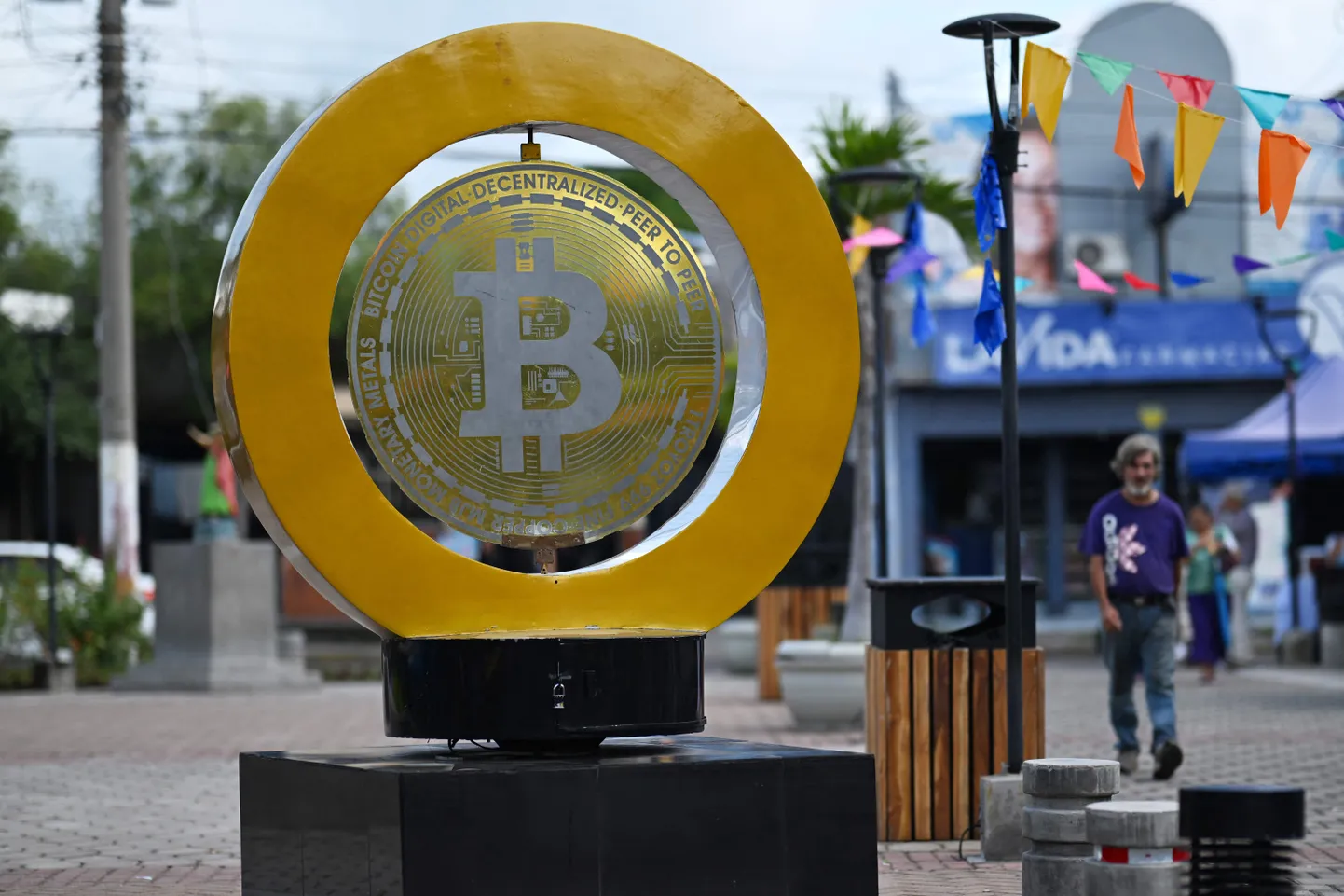 Mees kõnnib mööda bitcoin'i monumendist Plaza Bitcoinil San Salvadoris 4. septembril 2024. Avaliku arvamuse instituudi (IUDOP) andmetel on bitcoin'i kasutuselevõtt El Salvadoris endiselt piiratud hoolimata sellest, et see on olnud seaduslik valuuta juba kolmandat aastat. MTÜ Minu Esimene Bitcoin eesmärk on edendada krüptoraha kasutamist, õpetades seda riiklikes koolides, kus nende tundides on osalenud üle 35 000 õpilase. Kuigi 2023. aastal teatas 88 inimest 100st, et nad ei kasuta bitcoin'i, on mõned, nagu taksojuht Napoleon Osorio, seda oma ettevõtluses omaks võtnud.