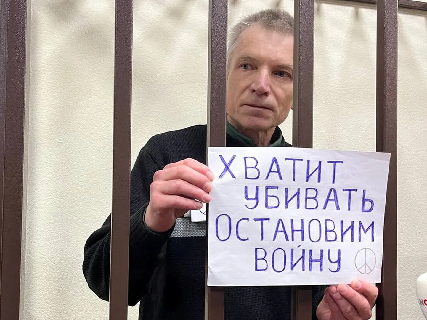 Алексей Горинов, отбывающий семилетний срок за критику вторжения России в Украину, держит плакат с надписью «Хватит убивать. Остановим войну», стоя в клетке для подсудимых во время нового судебного заседания против него за «оправдание терроризма» в разговорах с сокамерниками, Россия, 27 ноября 2024 года.
