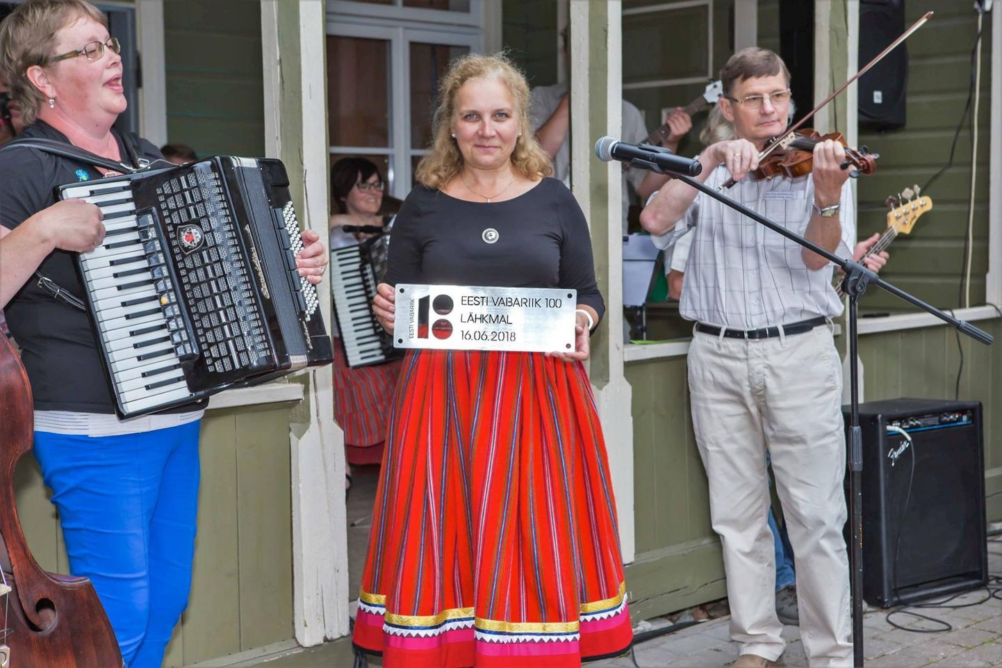 Lähkma-Saunametsa külaseltsi juhatuse liige Kadri-Aija Viik hoiab käes EV 100 juubeliplaati, akordioni mängib Õnnela Teearu ja viiulit Jaanus Männik.
