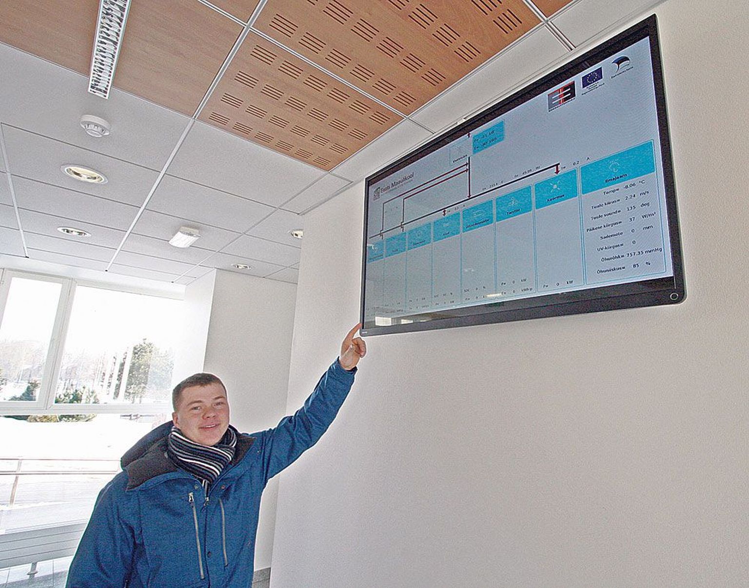 Maaülikooli energiakasutuse eriala doktorant Maido Märss näitab tehnikamaja fuajees paiknevat ekraani, millel jooksevad ilmaandmed ning taastuvenergia tootmise andmed reaalajas.