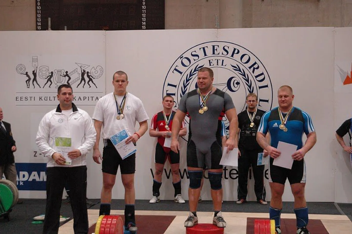 Eesti meistrivõistlused klassikalises tõstmises. Hetk asutasutamiselt. Esiplaanil vasakul treener Igor Baškirov. Pjedestaalil Lydon Mürgimäe, Lauri Nämi ja Erik Kuningas. Tagaplaanil Peeter Tammoja (punases) ja Riho Kägo.