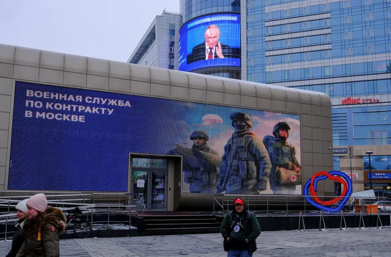 На здании реклама с надписью «Военнам служба по контракту в Москве», за ним, на другом здании, экран с лицом Путина.