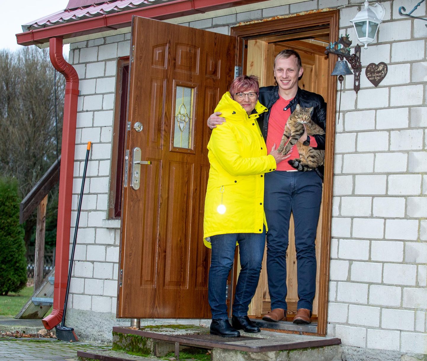 Elve ja Virgo Vahenõmm koos modellkassiga koduuksel, mille kõrval ripub südame­kujuline kiri “Tere tulemast meie koju!”.