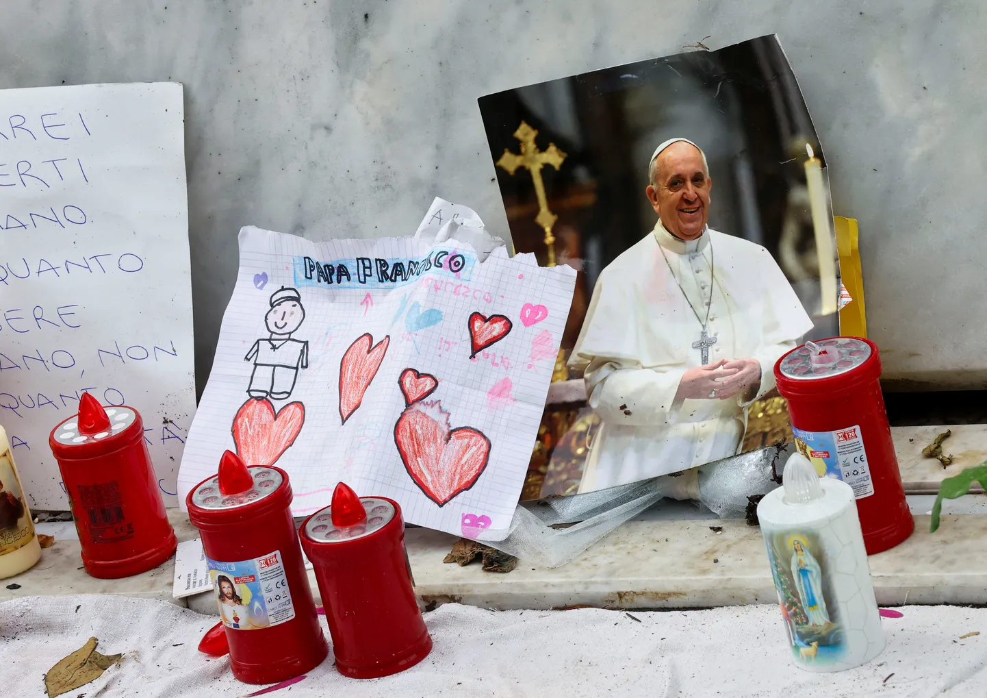 Pāvesta Franciska fotogrāfija blakus svecēm un zīmējumiem pie mirušā pāvesta Jāņa Pāvila II statujas pamatnes. Pāvests Francisks turpina ārstēšanos Gemelli slimnīcā Romā, Itālijā, 2025. gada 15. martā.